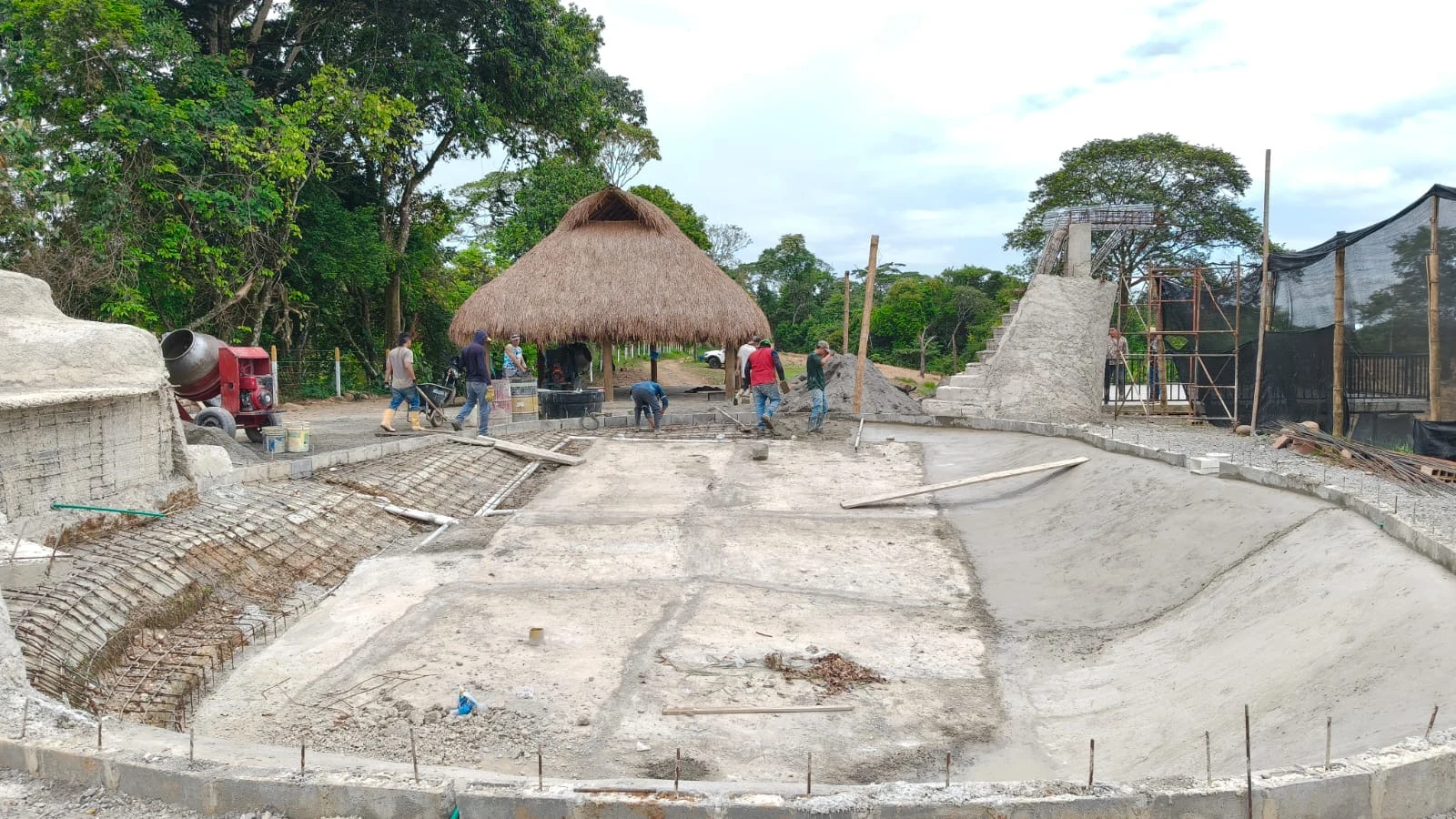 Piscina tipo playa en construcción, con obreros trabajando y kiosco de palma al fondo